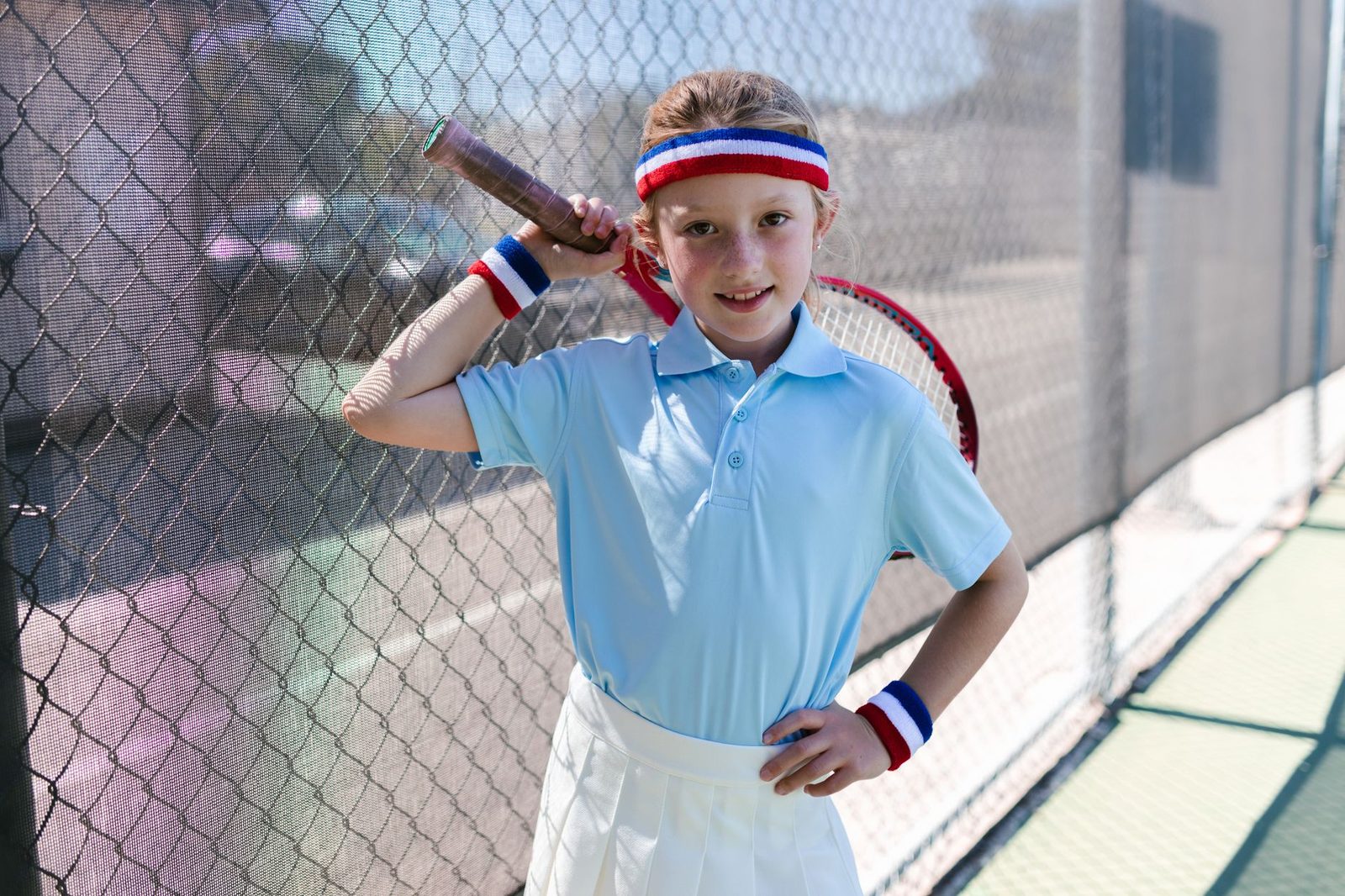 A smiling girl in tennis clothing standing by a fence with a racquet