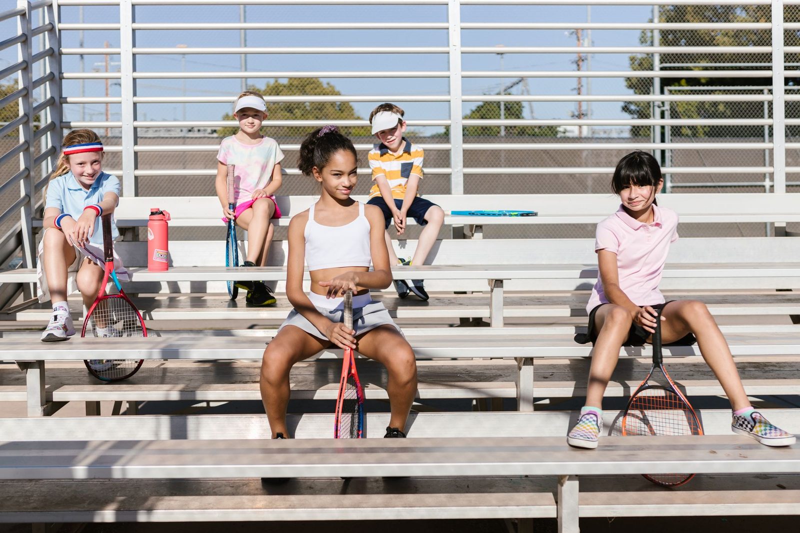 A group of children in sportswear sitting with tennis racquets on bleachers