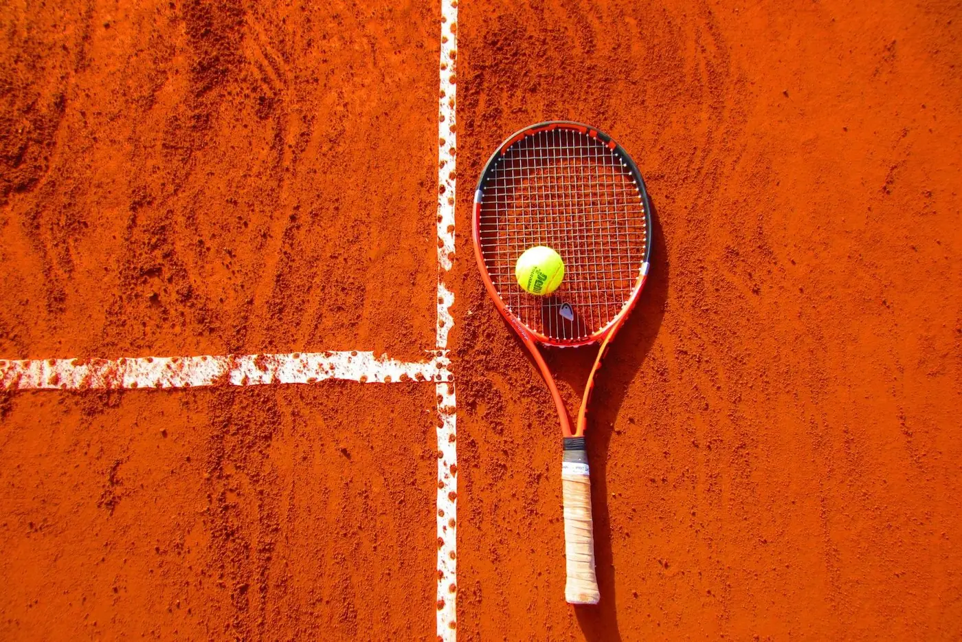 Freshly strung tennis racquet and ball resting on the line of a red clay court