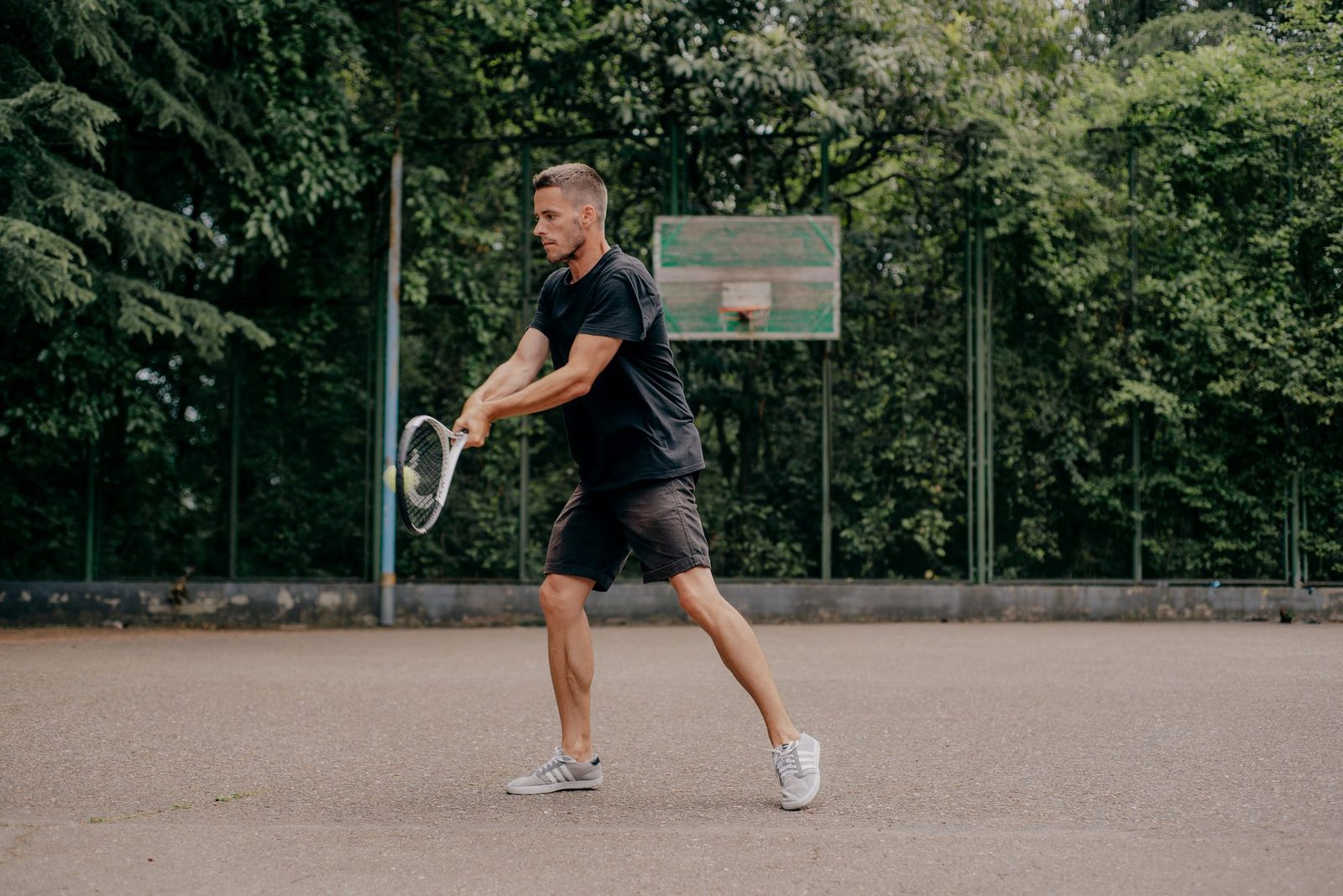 A young athlete playing tennis outdoors with trees behind the court
