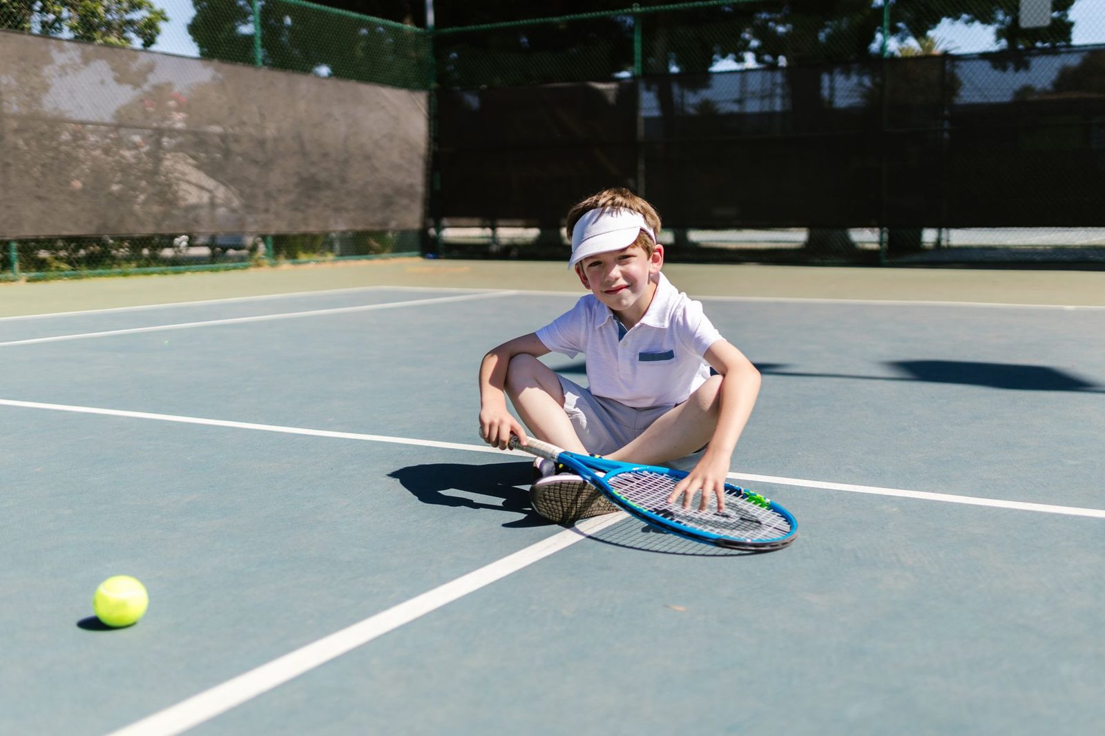 A cheerful young boy sitting on a tennis court with racquet and ball