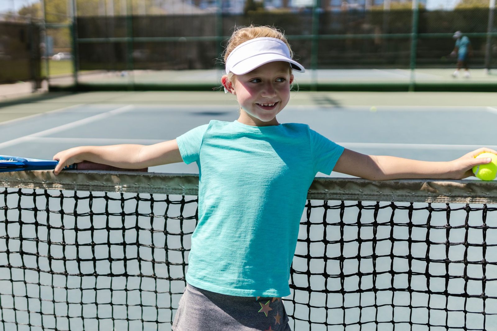 Smiling young girl in activewear leaning on a tennis net on a sunny day