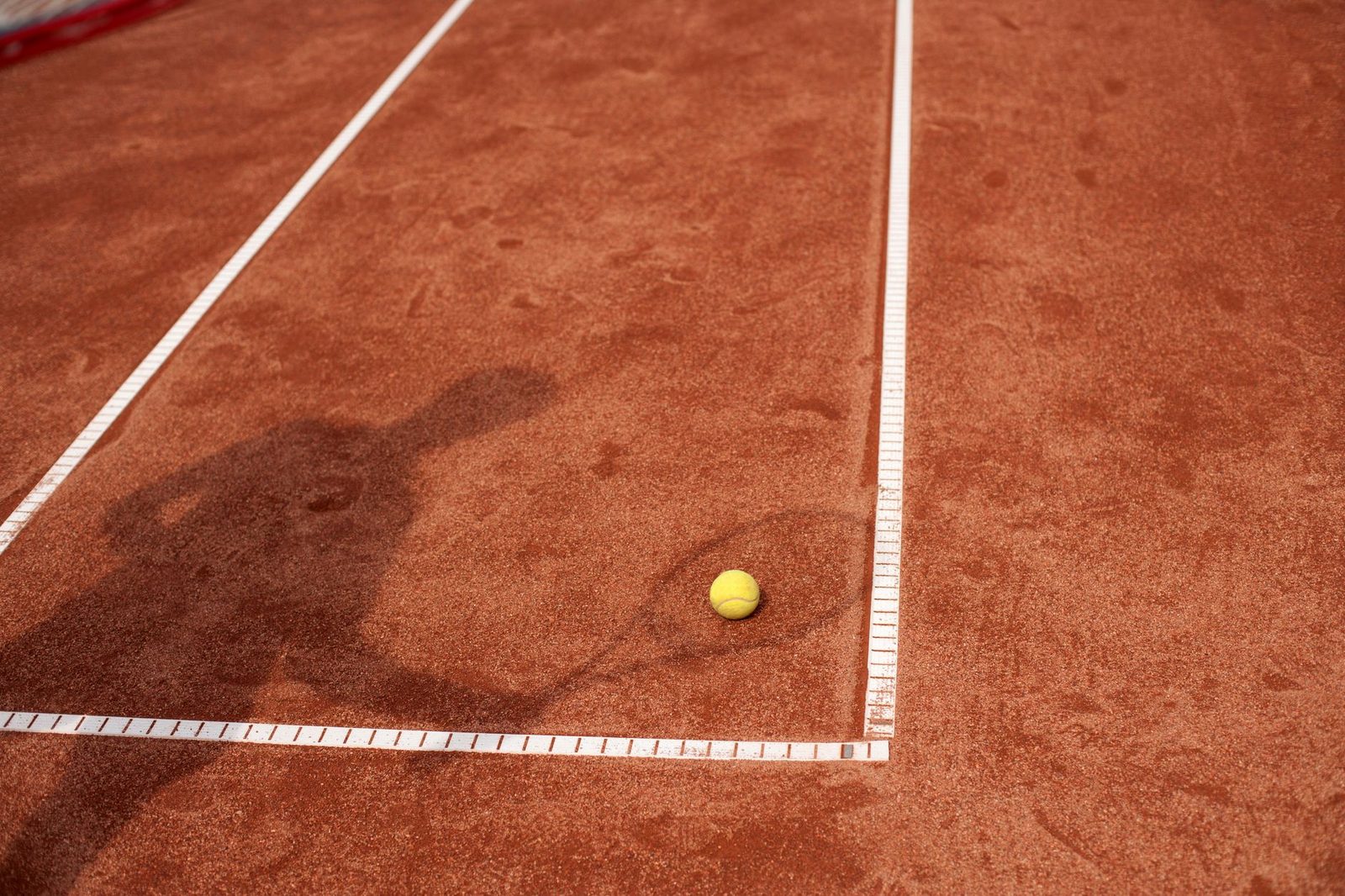Shadow of a tennis player on a clay court beside a yellow ball