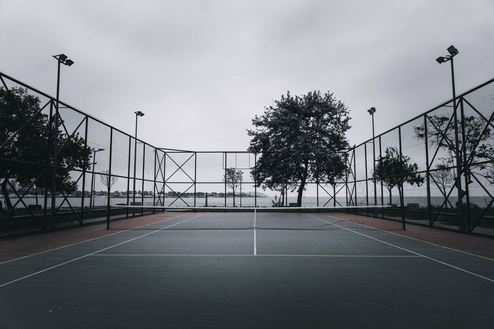 Empty outdoor tennis court enclosed by mesh fencing and surrounded by trees