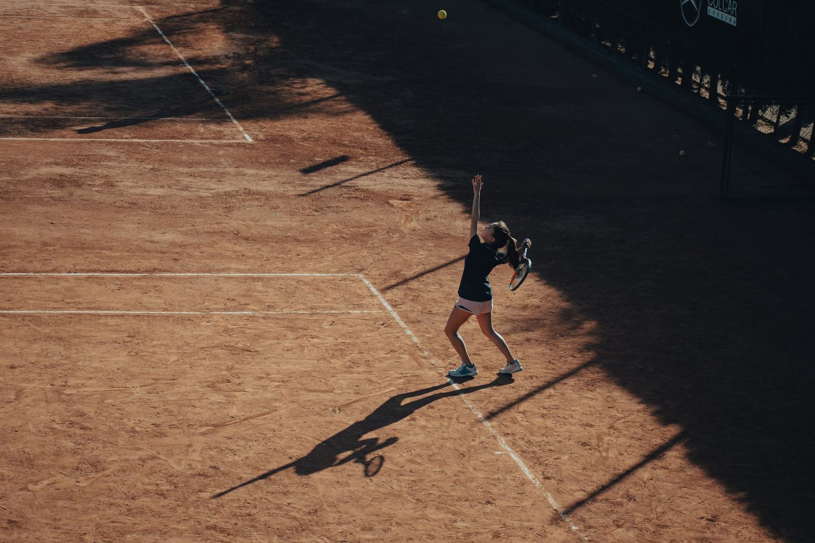 Aerial view of a woman serving on a sunlit clay tennis court