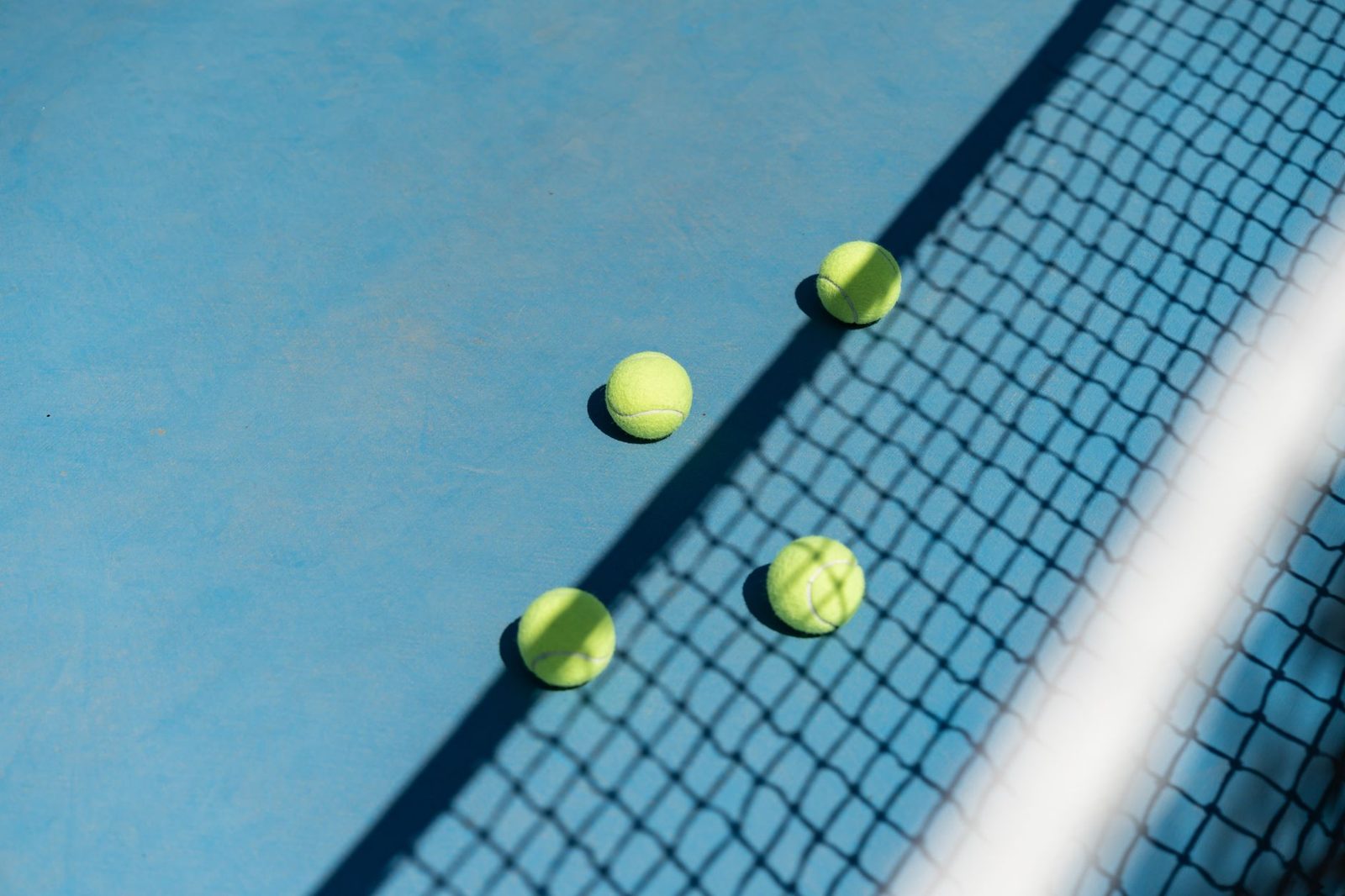 Three tennis balls scattered on a blue court beside the net shadow