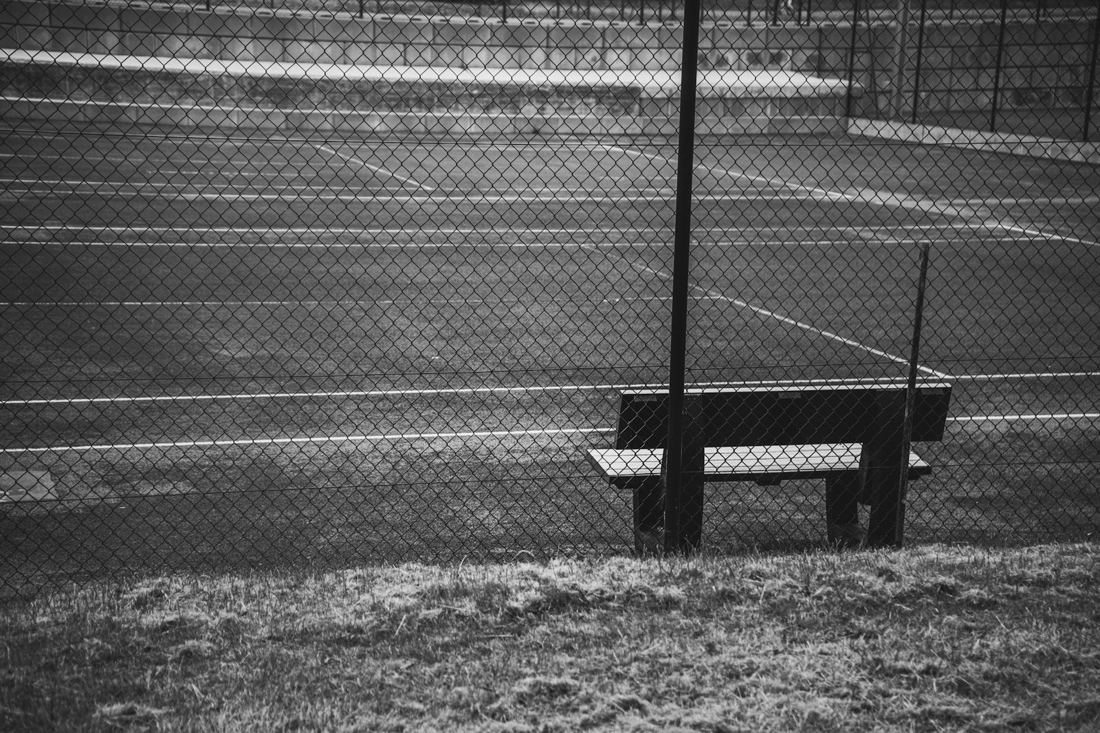 Empty tennis court and bench behind a chain-link fence, in black and white