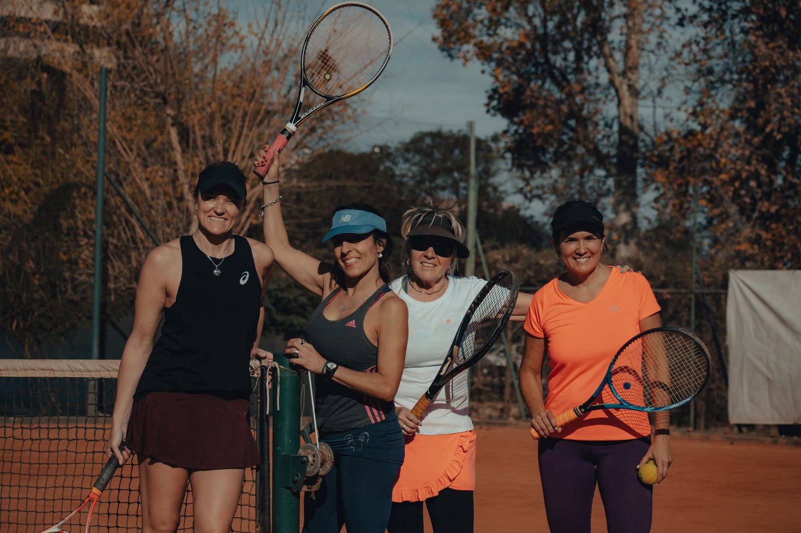 Four women holding tennis racquets and smiling together on an outdoor court