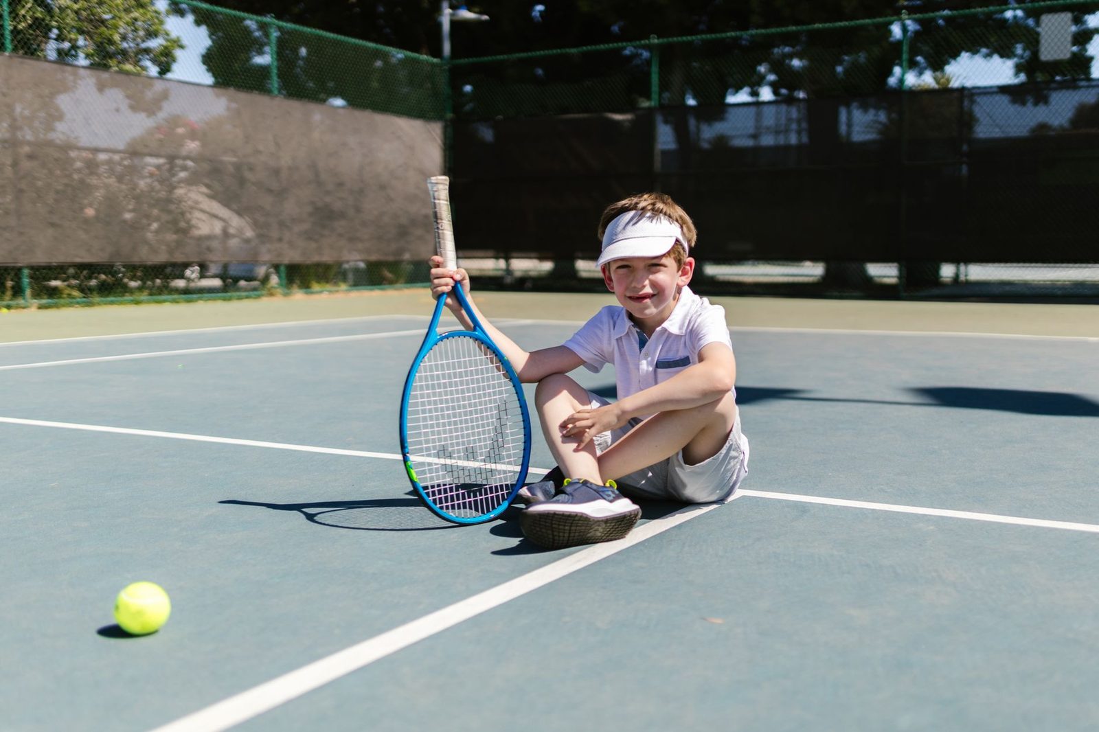 A cheerful boy sitting on a tennis court with a racquet and ball