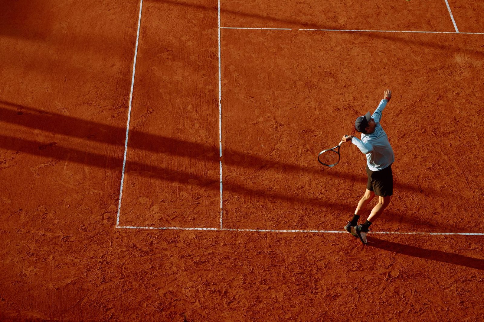 Player serving on a clay court at sunset, following through into the court