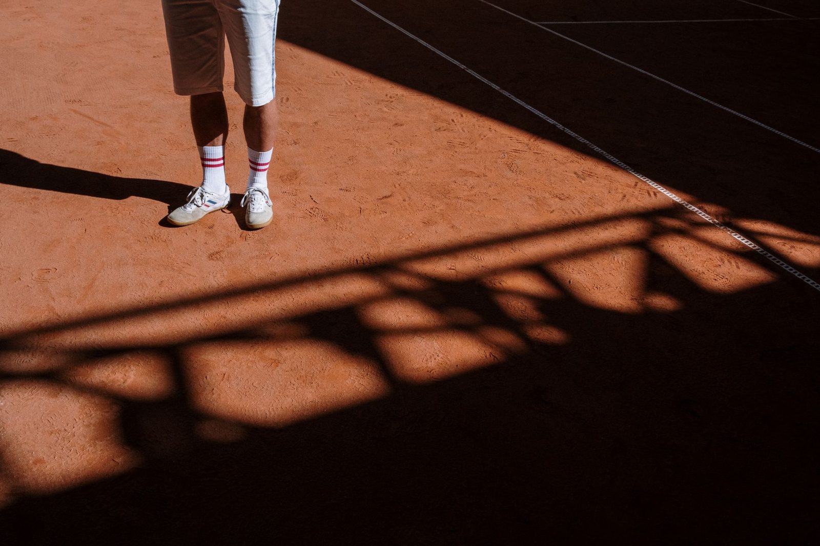 An athlete's legs and shoes casting a shadow on a sunlit clay court