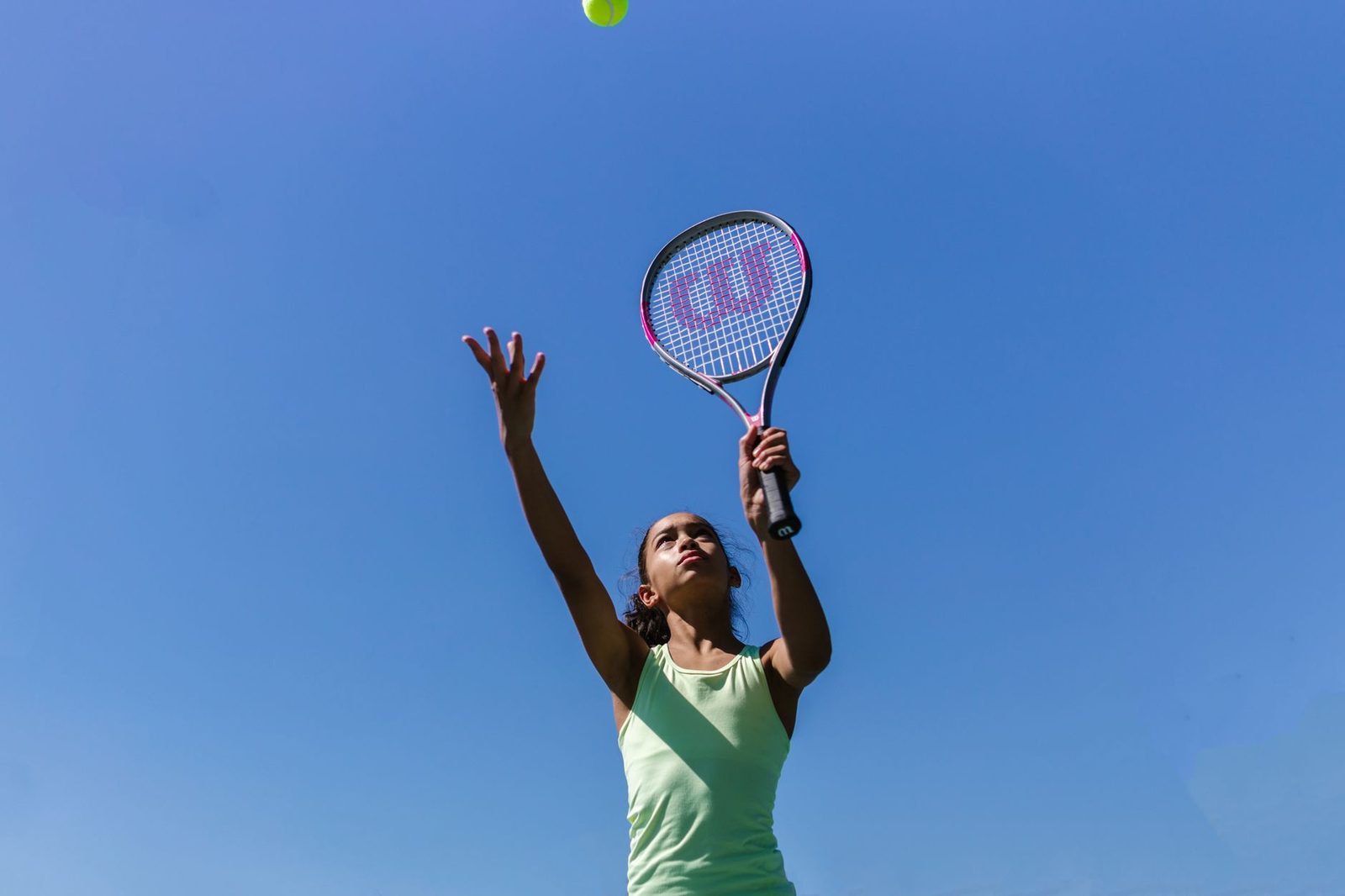 A teenage girl serving on an outdoor court under a blue sky